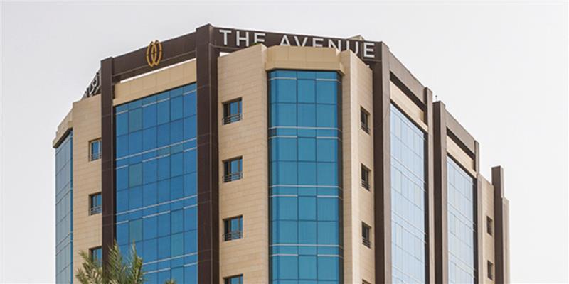 Modern multi-story office building with blue glass windows, beige stone accents, and The Avenue signage at the top.